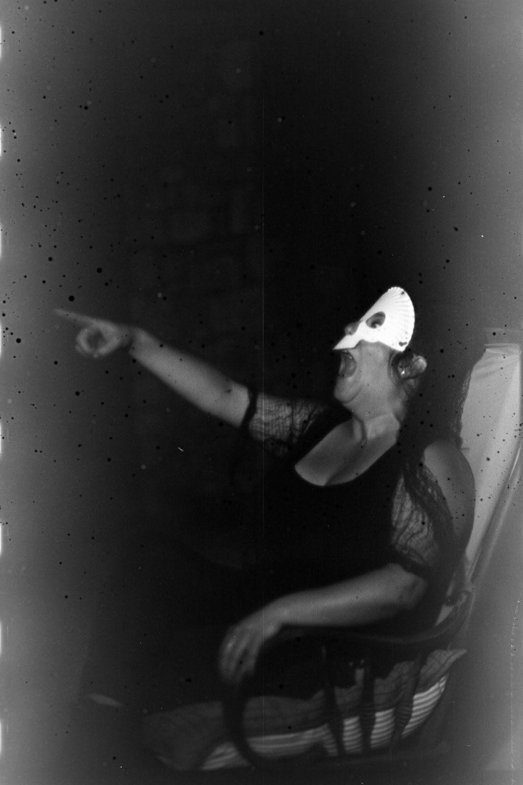 A person wearing a white mask and dark clothing sits in a chair, laughing and pointing to the side. The image has a grainy, vintage, black-and-white appearance.