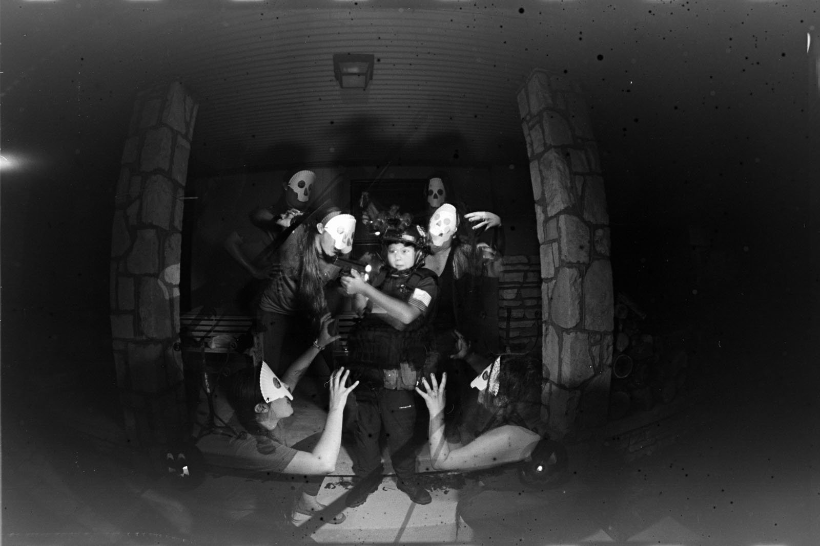 A person dressed as a police officer stands surrounded by figures in white masks and dark clothing, reaching toward them with outstretched arms, creating a haunting, ghostly effect in a dimly lit, eerie setting.