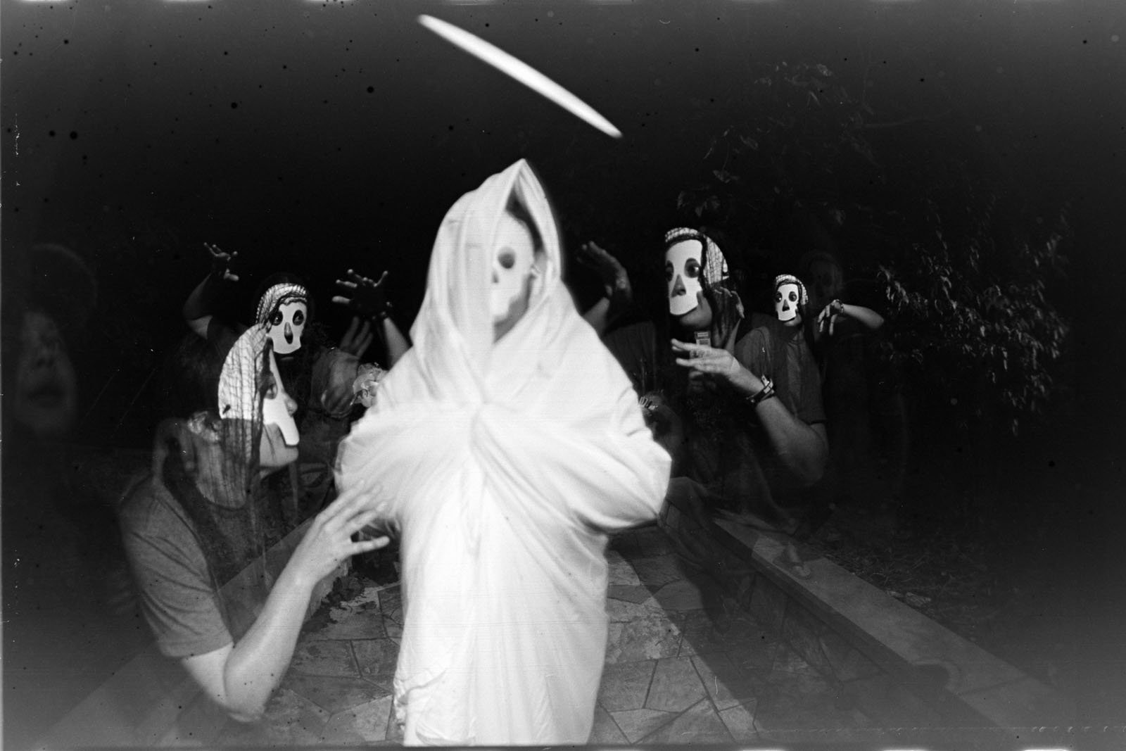 A person draped in a white sheet stands in the center, appearing ghostlike, surrounded by figures wearing white skull masks, all reaching toward the center. The image is black and white with an eerie, surreal atmosphere.