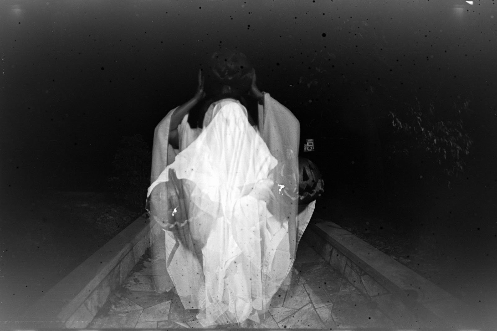 A person draped in a white sheet resembling a ghost stands on a stone pathway at night, holding a pumpkin. The image has a double exposure effect and black specks, creating a ghostly, eerie atmosphere.