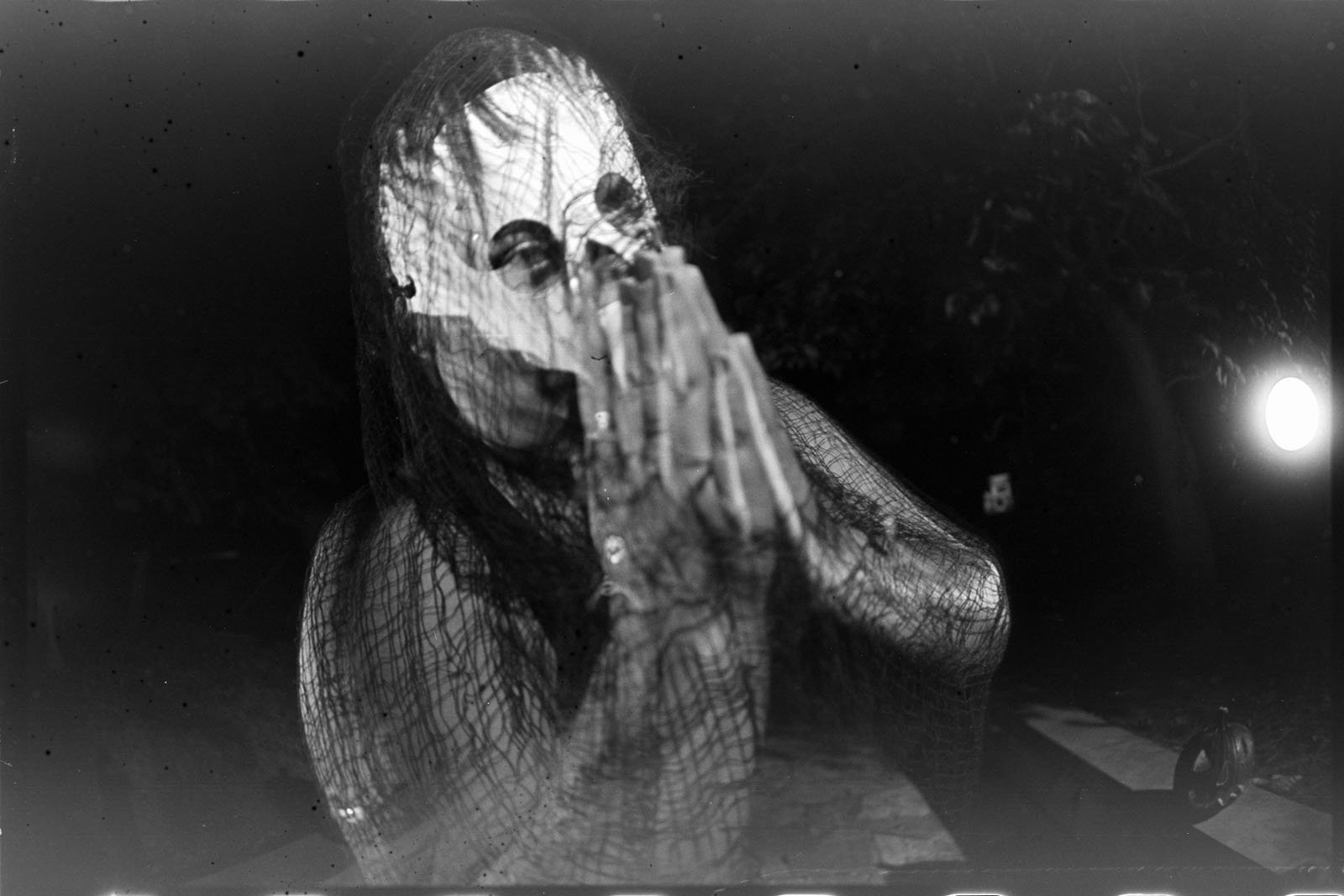 A person wearing a creepy, white mask and dark, netted clothing stands outdoors at night with hands pressed together in a prayer gesture. The scene is dimly lit, creating a haunting and mysterious atmosphere.