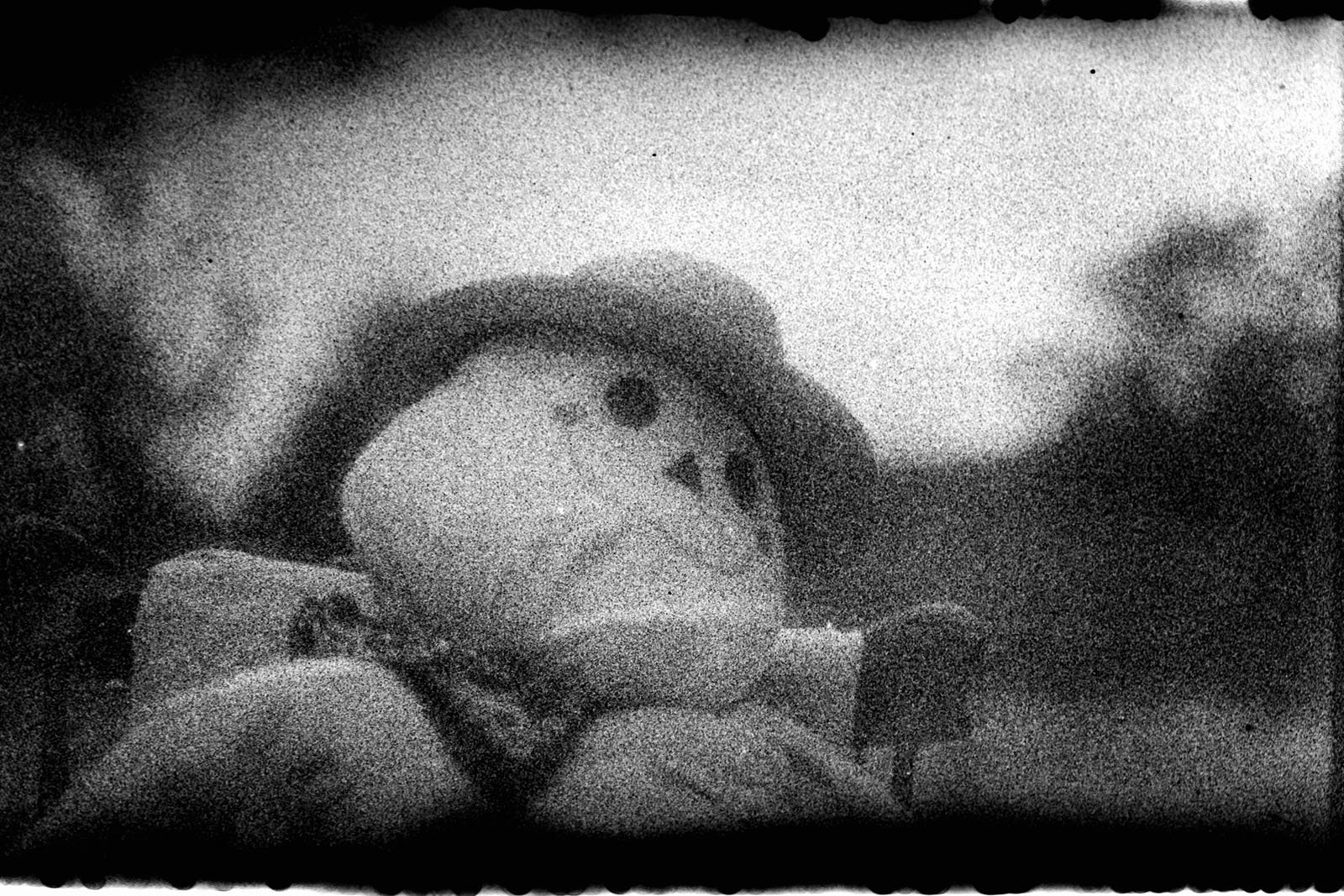 Grainy black and white photo of a stuffed toy with a round face, wearing a hat and bow tie, sitting outdoors with a blurry background and dark border edges.