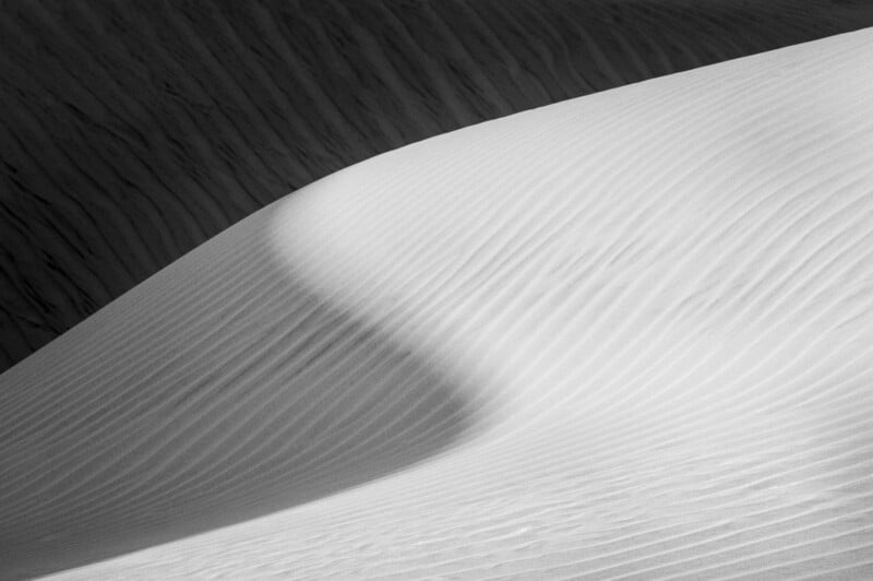 Smooth, white sand dunes with textured, wavy lines cast soft shadows, creating a flowing, abstract pattern in the sunlight. The image emphasizes the curves and natural beauty of the desert landscape.