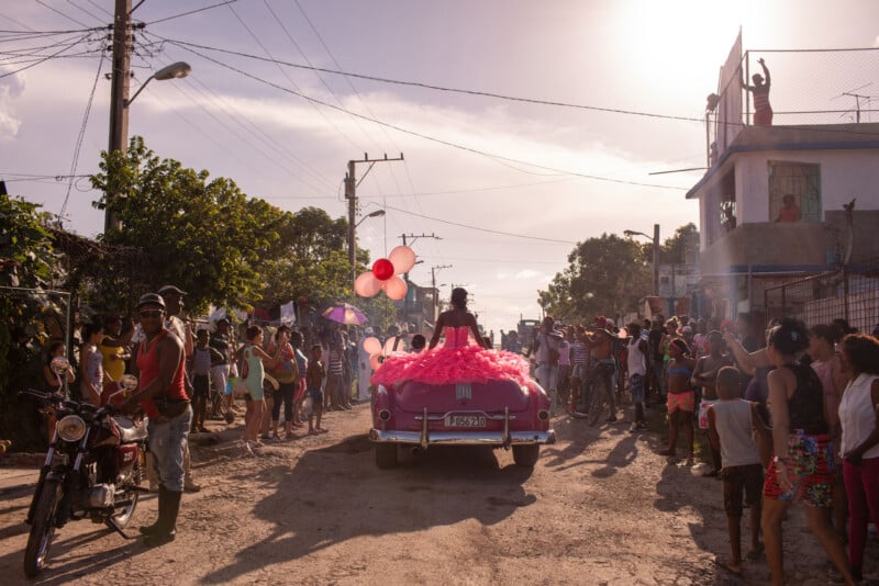 A vibrant street scene shows a crowd watching a classic pink car decorated with balloons and a girl in a dress riding on top, likely during a parade or celebration, with people taking photos and the sun setting in the background.