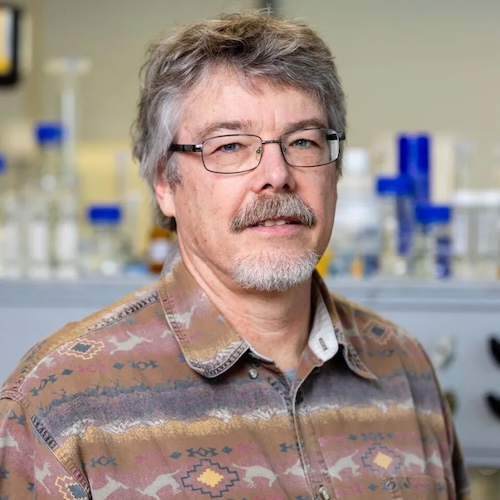 Man with grayish-brown hair, moustache and beard. Laboratory instruments are on a shelf in the background.