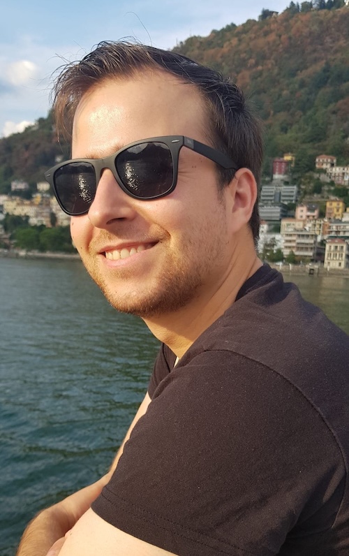 Smiling man at a waterfront wearing sunglasses and dark shirt.