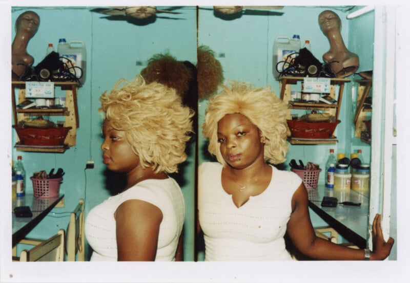 A woman with blonde hair stands beside a mirror in a salon, creating a double image of her. Behind her are shelves with hair products and wig stands, and the walls are painted light blue.