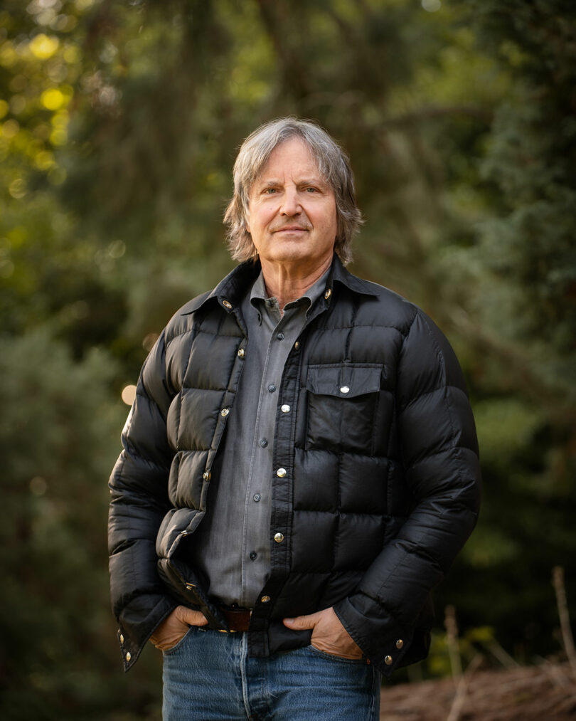 An older man with gray hair stands outdoors wearing a black puffer jacket and jeans, with trees in the background.