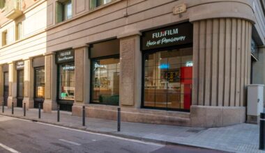 A corner view of a Fujifilm House of Photography store with large windows, black awnings, and a stone facade on a quiet city street. Bollards line the sidewalk in front of the shop.