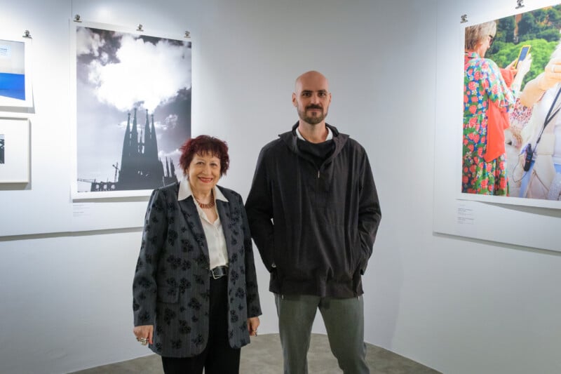 An older woman and a bald man stand in an art gallery, smiling at the camera. Framed photographs are displayed on the white walls behind them.