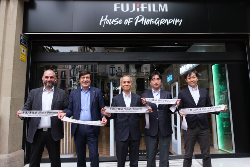 Five men in suits stand in front of a store named "FUJIFILM House of Photography," holding a ribbon with the store’s name, appearing to celebrate its opening. The storefront has glass doors and a black sign above.