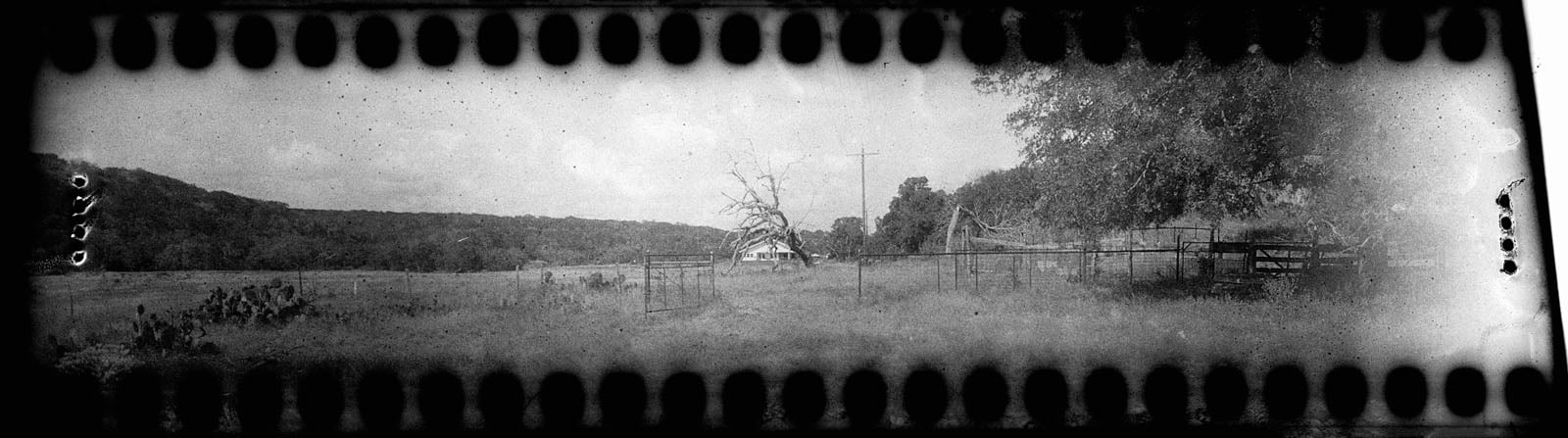 Black and white panoramic photo of a rural landscape with a fence, a gate, a tree, and a distant house. Hills are in the background. Film sprocket holes and edges are visible, giving a vintage effect.