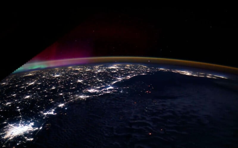 A view of Earth from space at night, showing city lights illuminating the surface below a dark sky, with a faint purple and orange glow along the horizon.