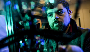 A bearded man in a dark hoodie gazes directly at the camera in a portrait lit with teal and blue lighting.