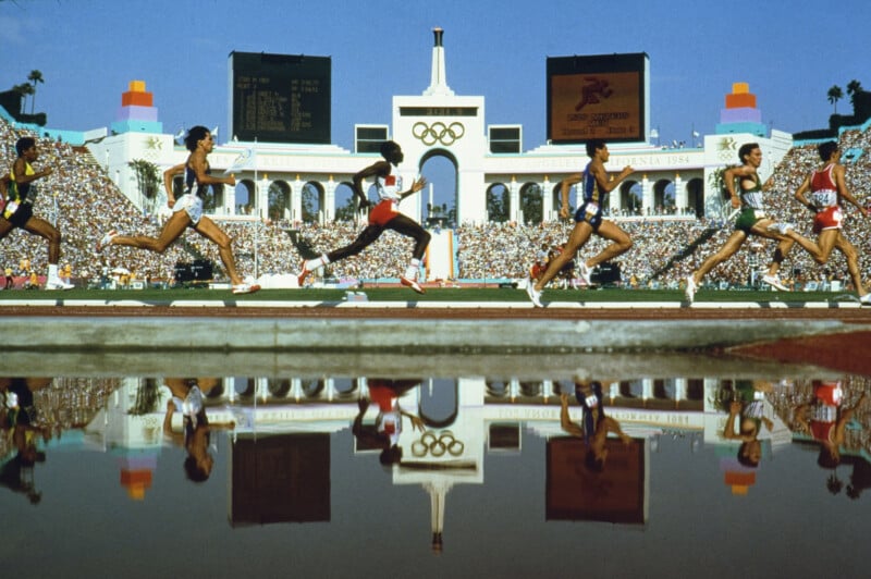 Runners compete on a track at an Olympic stadium with a large crowd in the stands. The iconic Olympic rings and columns are visible in the background, and the athletes are reflected in a pool of water in the foreground.