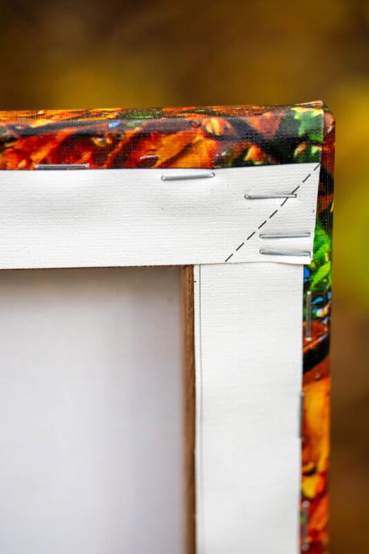 Close-up of the back corner of a stretched canvas painting, showing the wooden frame, stapled edges, and part of the colorful painted front wrapping around the corner. The background is blurred.