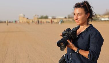 A woman wearing a dark shirt and sunglasses on her head stands on a wide, sandy road holding a camera. The background is blurred with people and buildings visible in the distance.