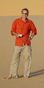 A man in a long-sleeve orange shirt and khaki pants standing in the desert holding a camera.