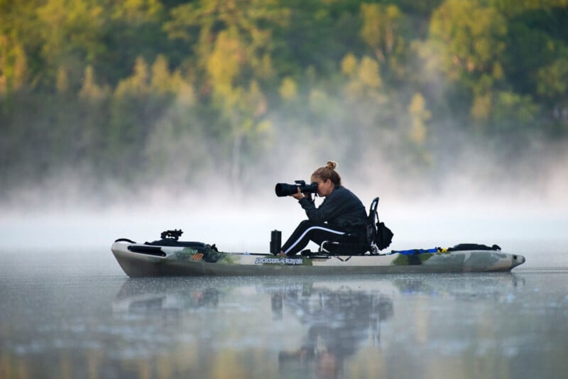 A person sits in a kayak on calm water, surrounded by mist, taking photos with a large camera. Trees and greenery are visible in the background.