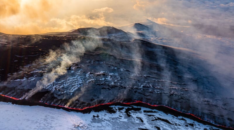 Aerial view of a volcanic landscape with a glowing line of lava, steam rising from the ground, patches of snow, and mountains in the background under a cloudy, golden sky.