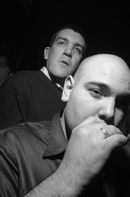Two men are standing close together in dramatic lighting. The man in the foreground is bald, wearing a ring, and has his hand near his mouth. The man in the background looks slightly upward with a serious expression.