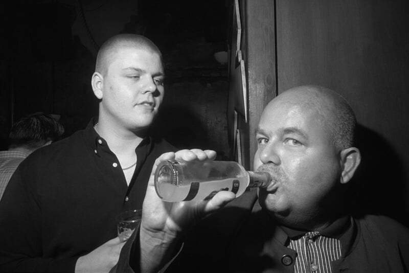 Two men in a dimly lit room; one is drinking from a bottle while the other, holding a glass, looks at him. Both have short hair and wear dark shirts. The atmosphere appears casual and social.