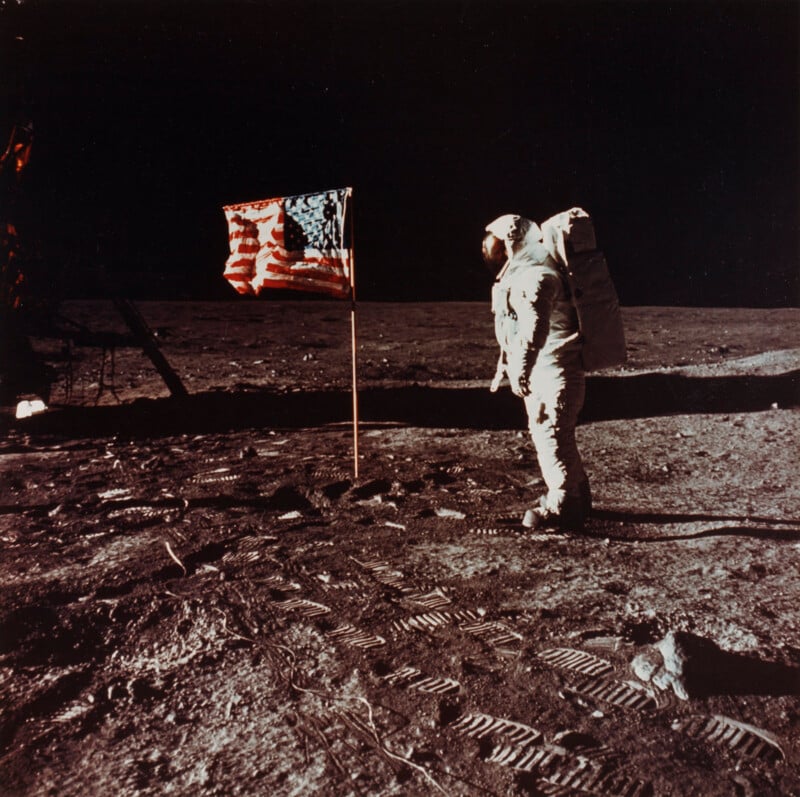 An astronaut in a spacesuit stands on the moon’s surface near an American flag, with numerous footprints and the lunar lander visible under a dark sky.