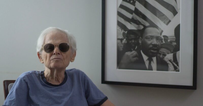 An older person wearing sunglasses and a blue shirt sits next to a wall with a framed black-and-white photo of Martin Luther King Jr. holding his fist up, with American flags visible in the background.