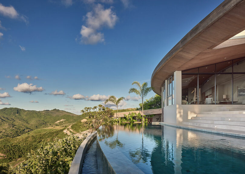 Modern villa with floor-to-ceiling windows overlooks a mountain landscape, featuring an infinity pool and clear blue sky.