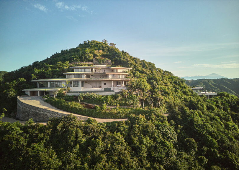 Large modern house with spacious balconies situated on a lush, green hillside under a clear sky, with a winding driveway and distant mountains in the background.