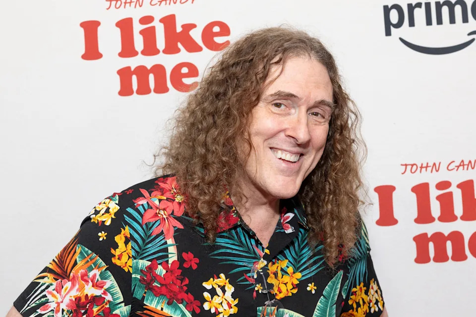 Person with curly hair in a floral shirt smiling at a "I Like Me" event backdrop