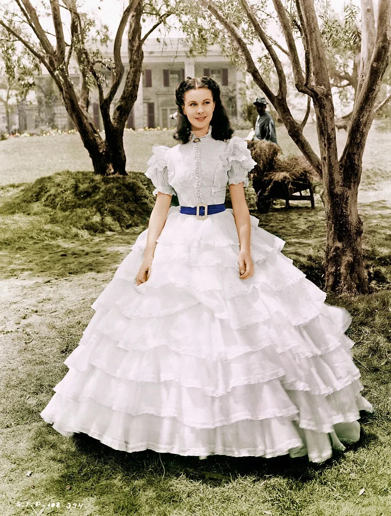 Leigh in period-style gown with ruffled layers poses outdoors, with trees and a building in the background