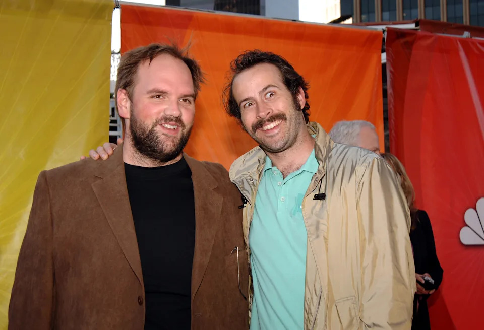 Two men smiling at an event, one in a brown blazer and black shirt, the other in a casual beige jacket and light shirt. Background features colorful panels