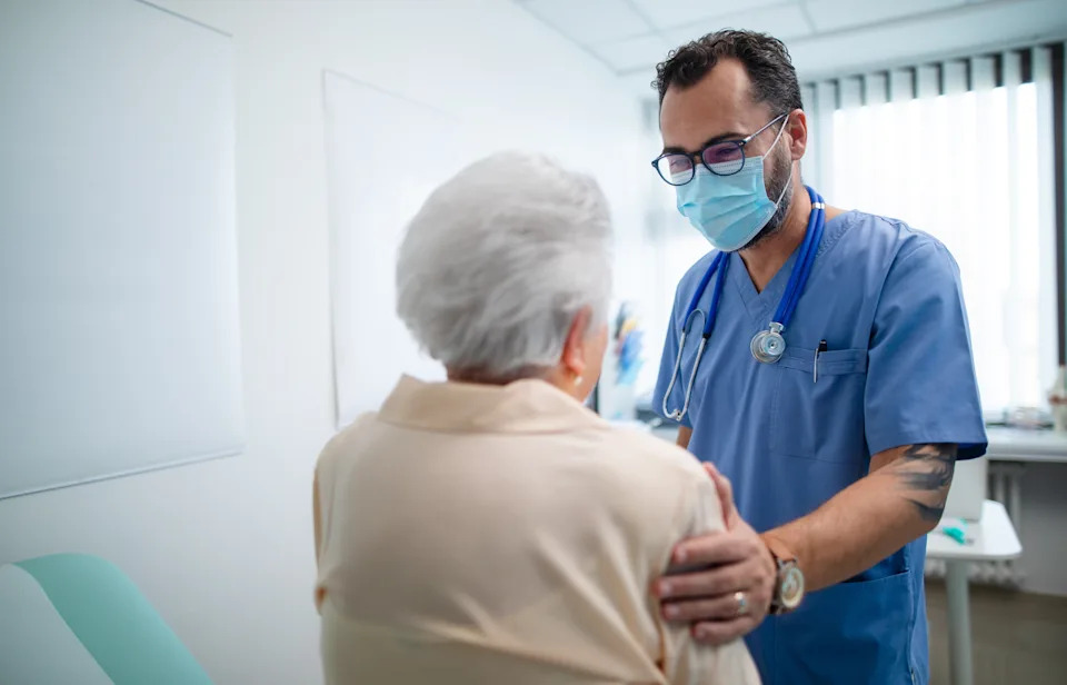 A healthcare professional in scrubs and a mask converses with an elderly patient seated in a clinic
