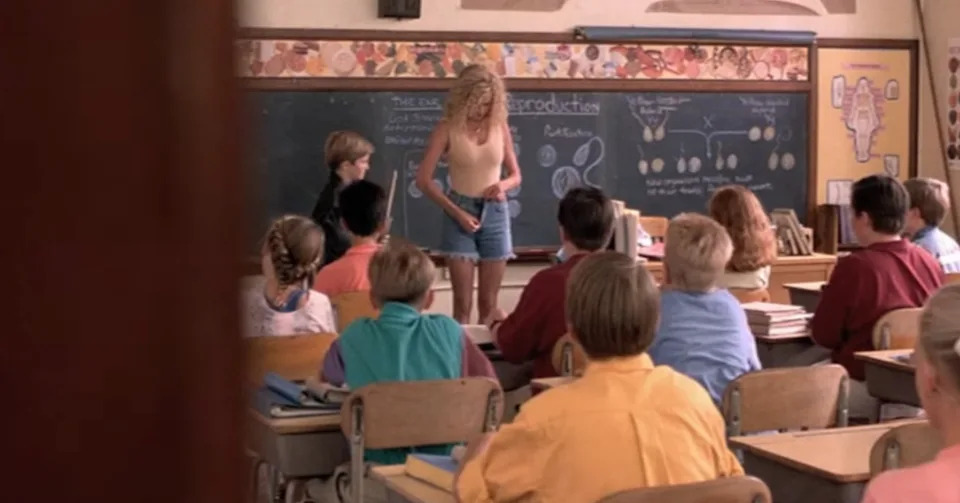 An adult woman stands at the front of a class unzipping her shorts