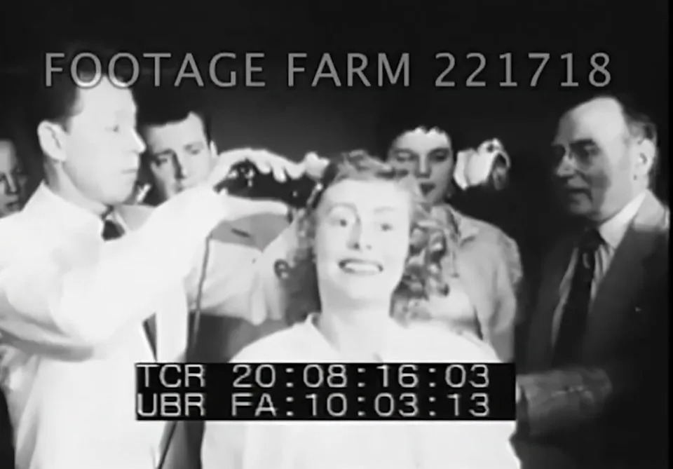 Smith smiles excitedly as surrounded by men operating a clipper over her head in a vintage setting