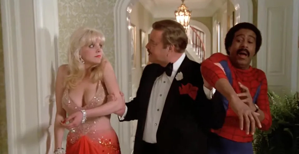 A man in a tuxedo guides a woman in a sequined gown; another man, dressed as Spider-Man, gestures animatedly
