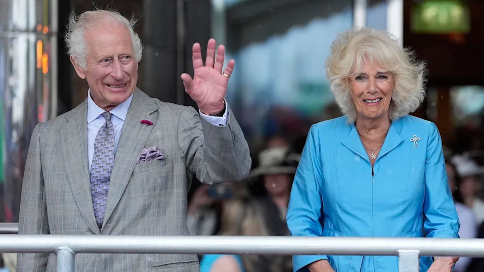 King Charles III and Queen Camilla stand on a platform while on a royal visit to Jersey. The King has a grey suit on and is waving to crowds with his left hand. The Queen has a sky blue dress on.