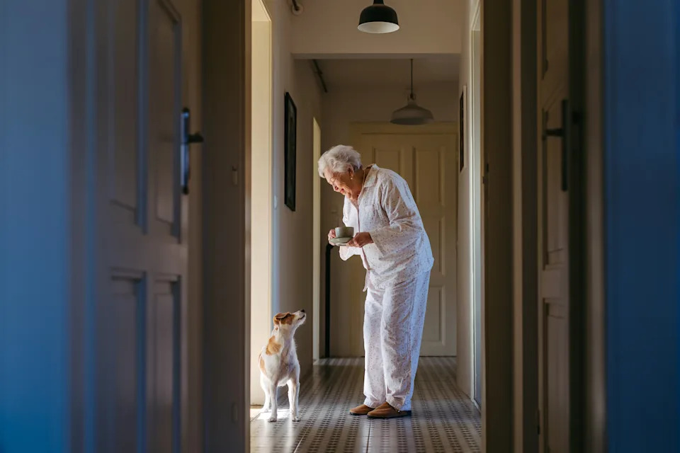 Elderly person in pajamas holding a bowl in a hallway, smiling down at a seated dog