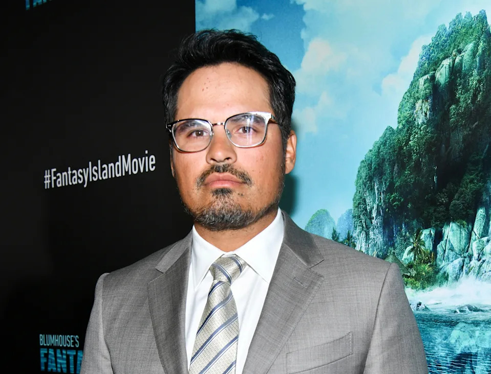 A person in a gray suit and striped tie at a movie premiere, standing in front of a poster with "Fantasy Island" hashtag visible