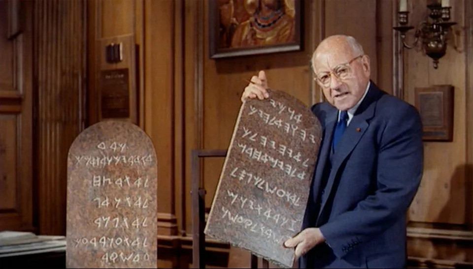 DeMille in a suit displays stone tablets with ancient script, resembling biblical Ten Commandments tablets, in a wood-paneled room