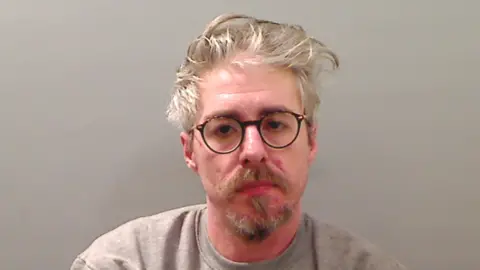 PSNI Mugshot of Barry Maguire. He has grey wavy hair and goatee, wearing glasses and a grey sweatshirt. 