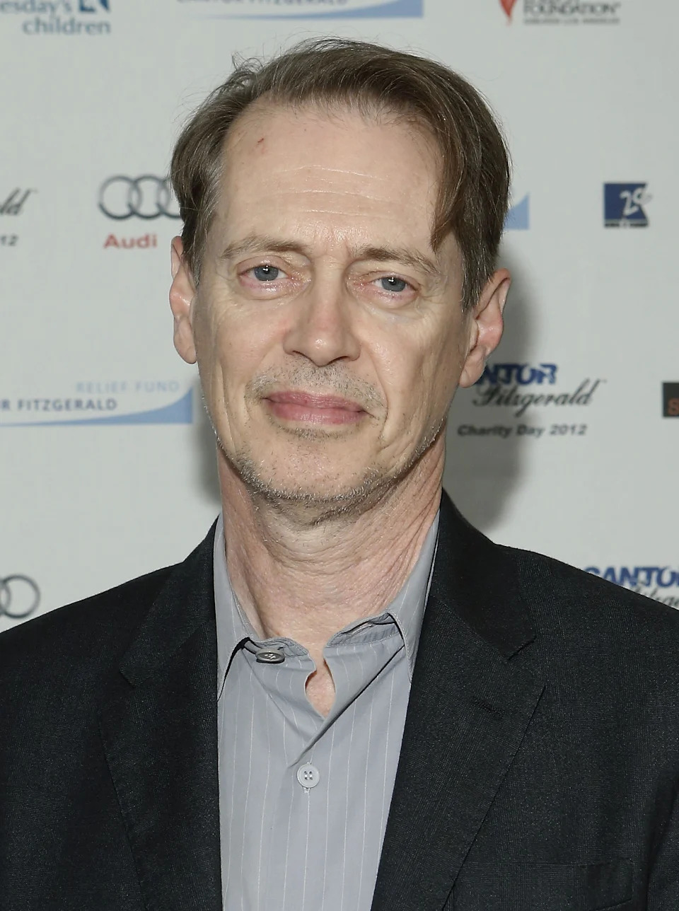 Steve Buschemi in a suit jacket and shirt at a formal event with branded backdrop, smiling slightly
