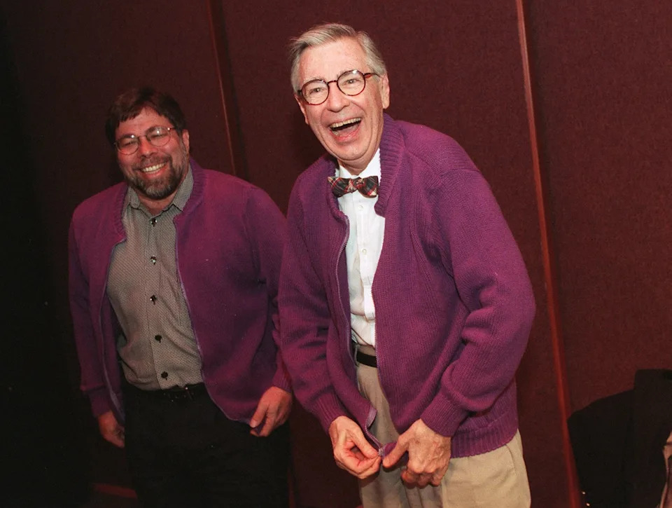 Two smiling people wearing similar cardigans, one with glasses and a bow tie, standing together in a casual setting