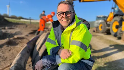 Professor McDonald kneels beside the skull of the fin whale at the dig site. He is wearing a hi-vis jacket and is smiling. There are workers behind him helping to steady the skull as it is placed on the ground.