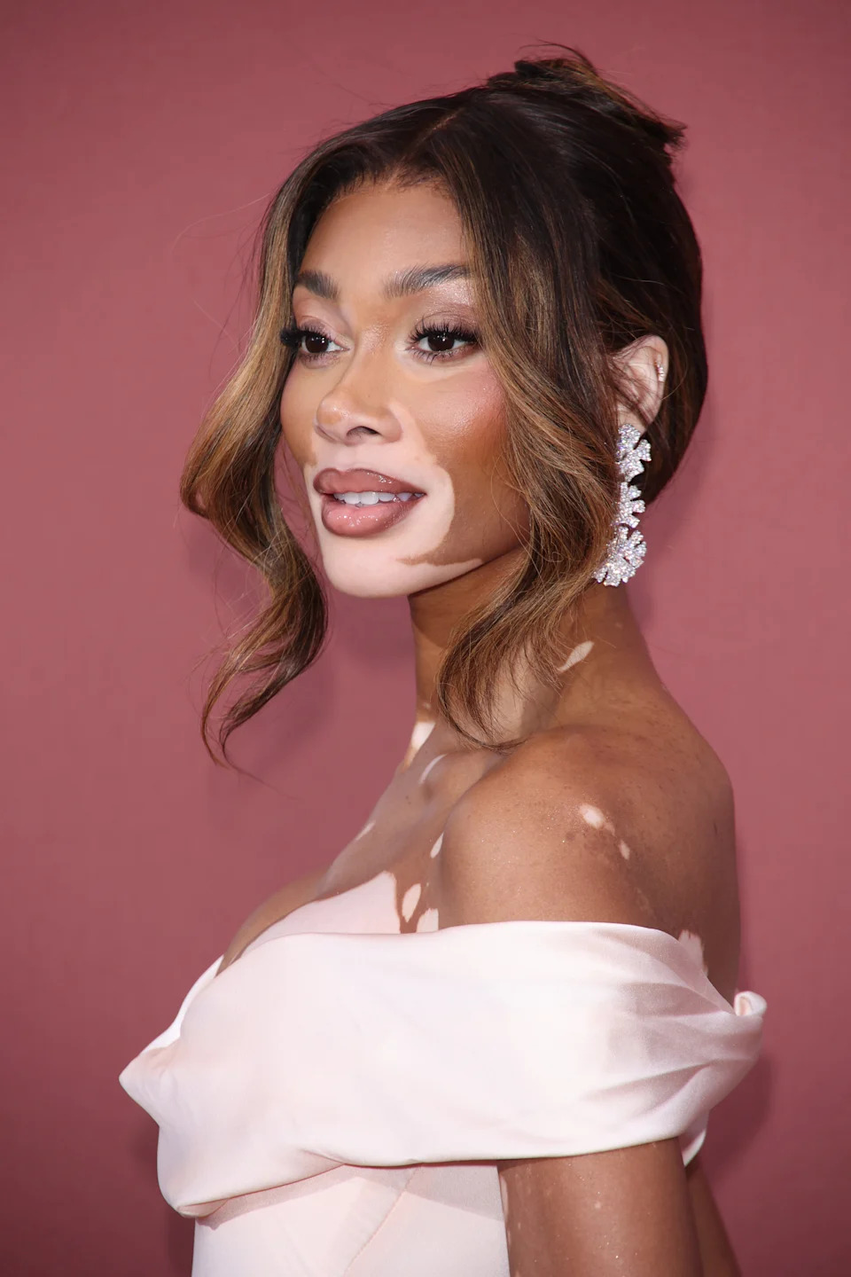 Winnie Harlow on red carpet wearing an elegant off-shoulder dress and statement earrings, looking to the side