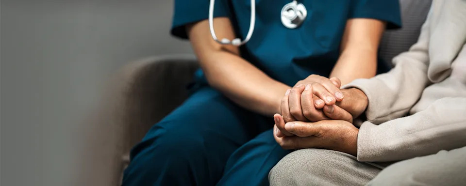 A healthcare worker in scrubs gently holds an older adult's hand, offering comfort and support