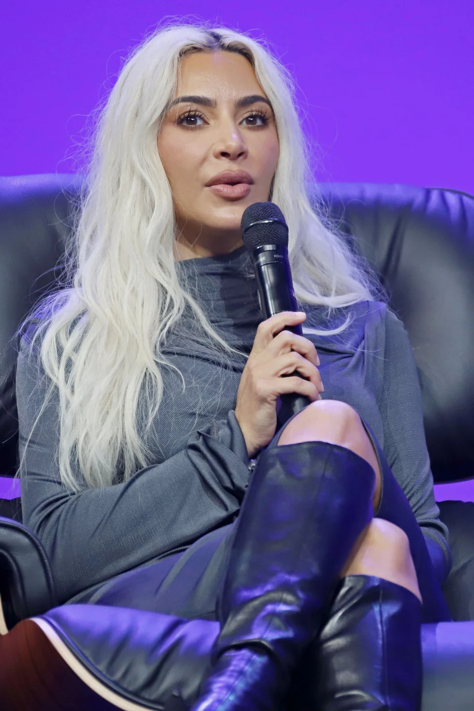 Kim Kardashian in a chic long-sleeve outfit and knee-high boots speaks into a microphone while seated on stage
