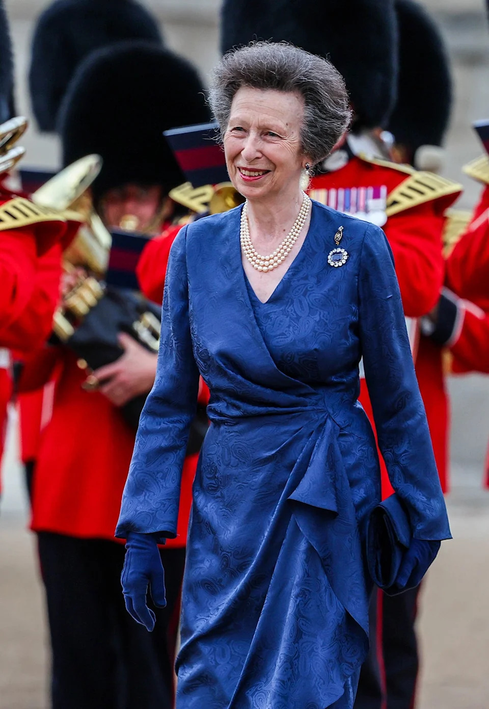 Princess Anne wearing a blue dress walking in front of British guards and smiling.