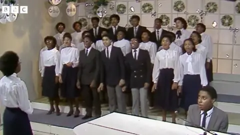 BBC Pebble Mill The women in the choir are wearing white blouses and skirts and the men, dark jackets and trousers and a white shirt.